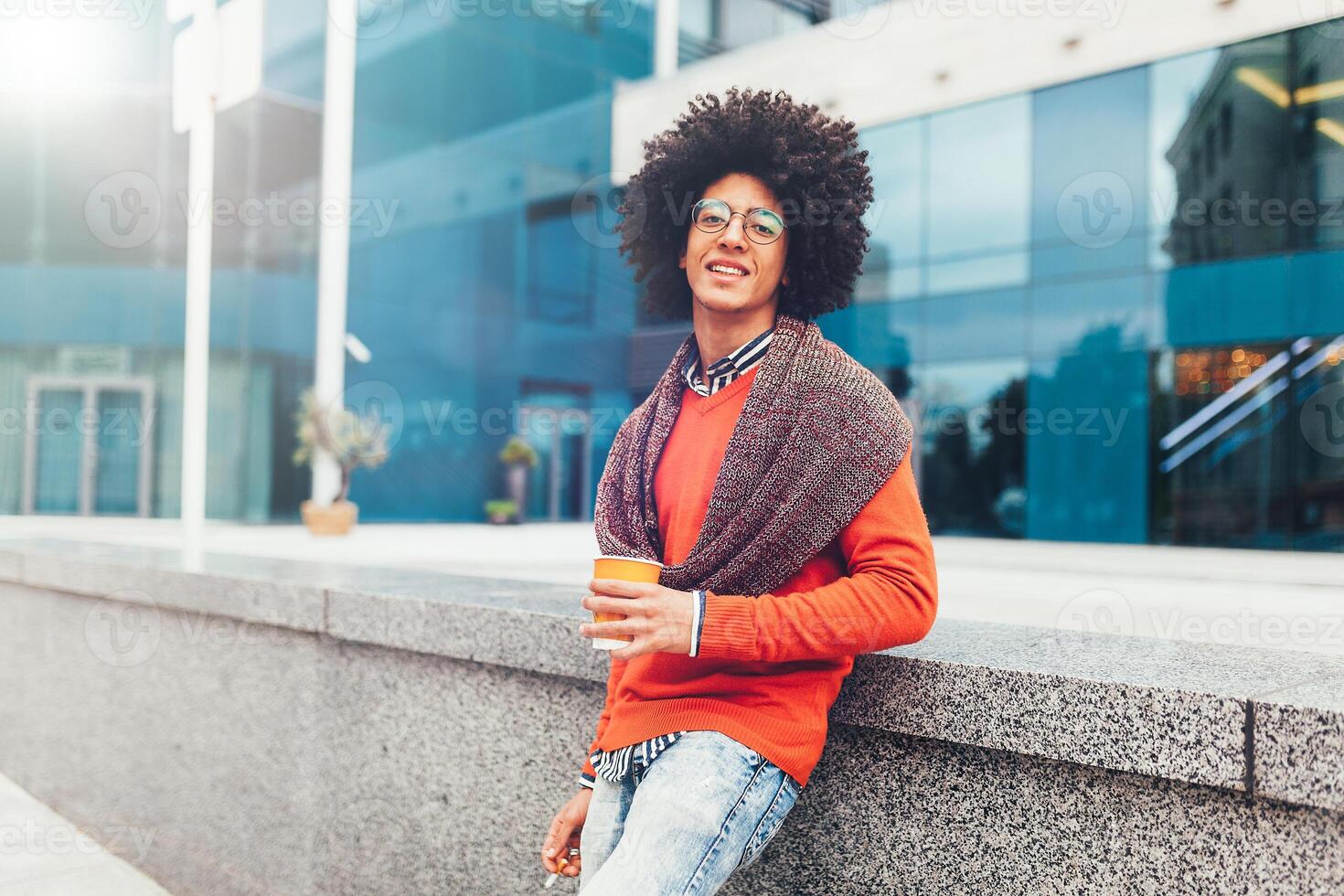 Young curly mixed-race boy drinks coffee on the street against the background of office buildings. Youth culture and fashion. Student photo