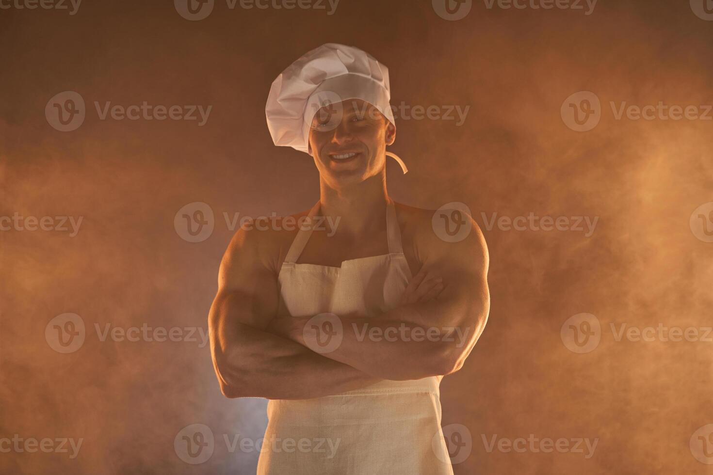 retrato de muscular cocinero vistiendo blanco delantal y cocinero sombrero, posando con doblada brazos en ahumado antecedentes foto