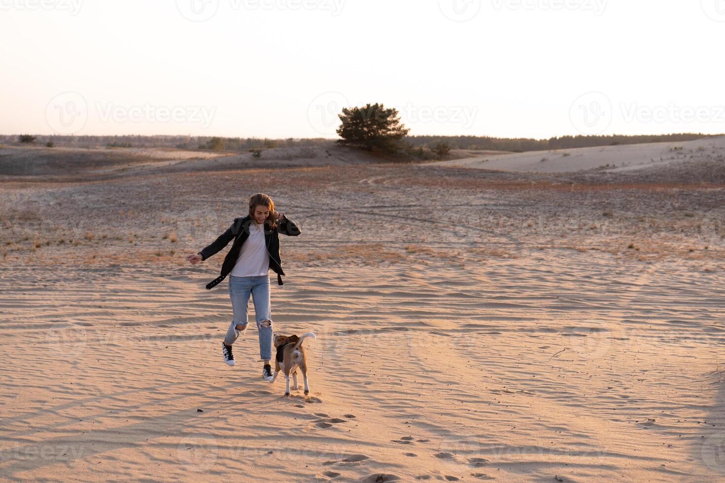 a young Caucasian girl somewhere in a leather jacket and blue jeans runs along the sandy beach with her beagle dog photo