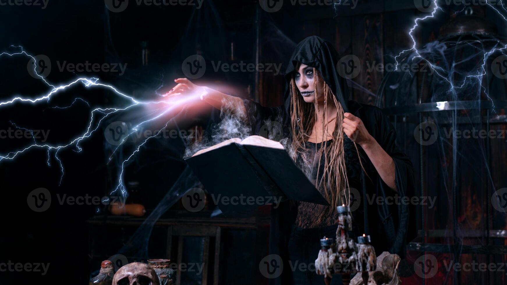 Halloween concept. Witch dressed black hood with dreadlocks standing dark dungeon room use magic book for conjuring magic spell photo