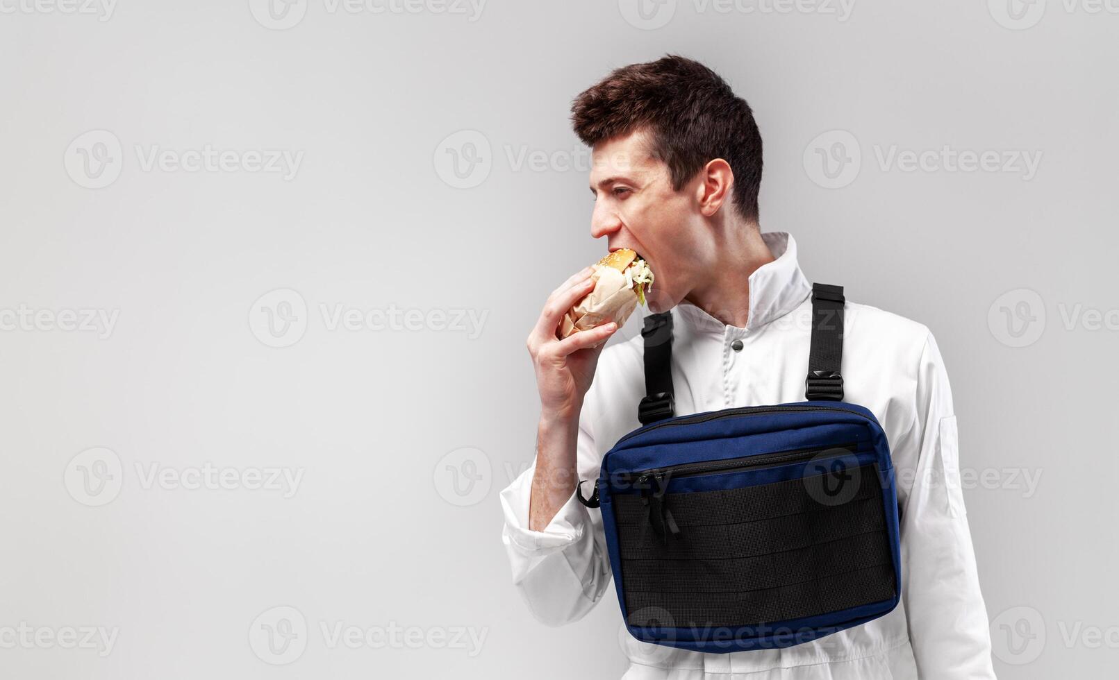 joven elegante masculino trabajador con cofre plataforma bolso es comiendo un sabroso jugoso hamburguesa en un blanco antecedentes foto