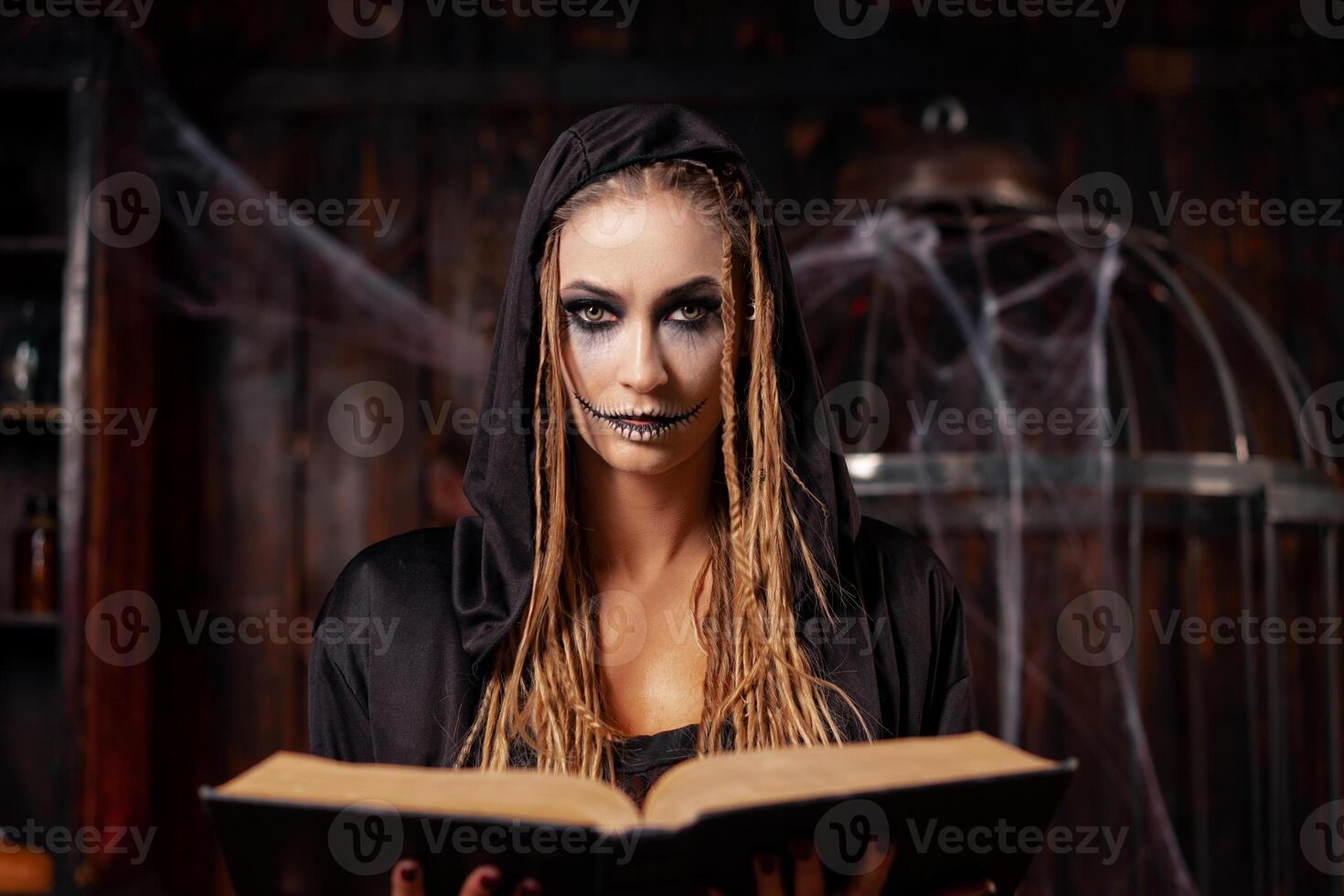 Halloween concept. Witch dressed black hood with dreadlocks standing dark dungeon room use magic book for conjuring magic spell photo