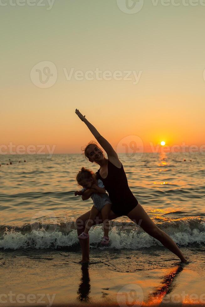 woman and girl silhouette practicing balancing yoga warrior pose together during ocean sunset with bright orange sky and water reflections photo