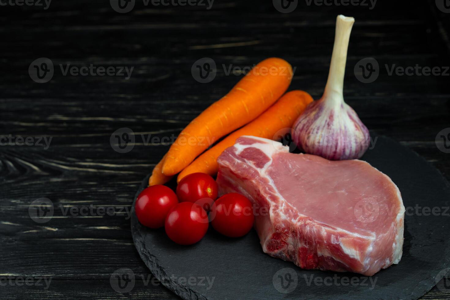Top view of one pieces raw pork chop steaks with cherry tomatoes carrot and garlic on a black stone cutting board. photo