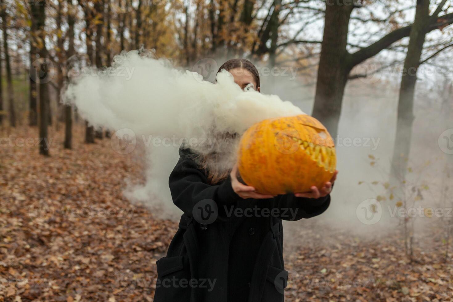 un hermosa niña con largo pelo en un negro capa es participación un naranja calabaza desde cuales fumar es próximo. contento Víspera de Todos los Santos. oscuro otoño bosque. horrores foto