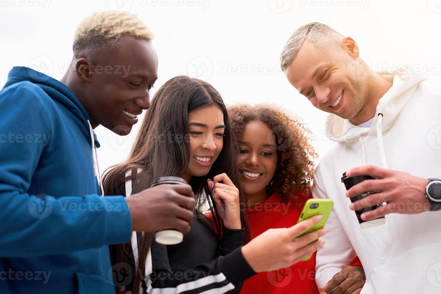 Young people, students of different nationalities, smile, hold coffee in their hand and look into one smartphone. Spend time together with friends. Two men and two women. White background. photo