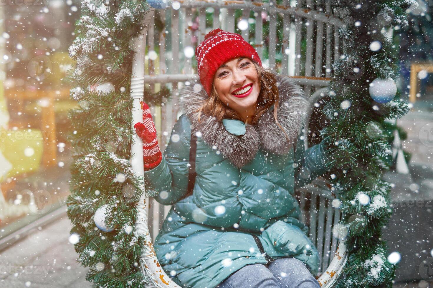 Young beautiful caucasian girl dressed in a warm winter jacket, knitted hat and gloves sits on Christmas decorated swing outdoor photo