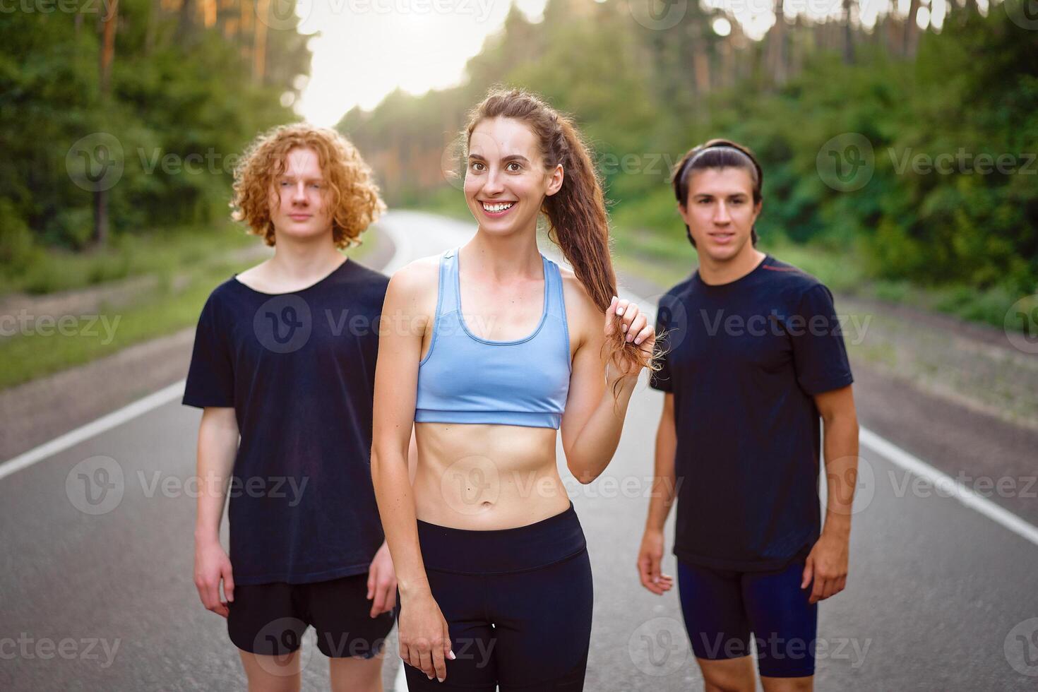 A group of three people athletes one girl and two men standing on asphalt road in pine forest at summer before jogging photo