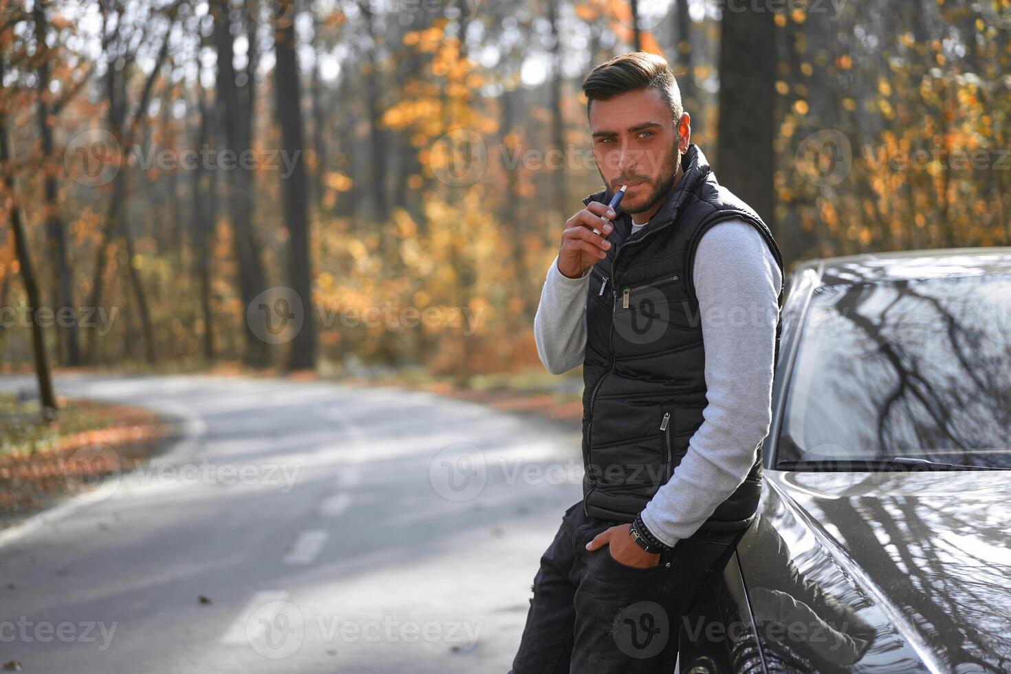 A handsome young Caucasian man wearing style black vest standing near black car on nature. photo