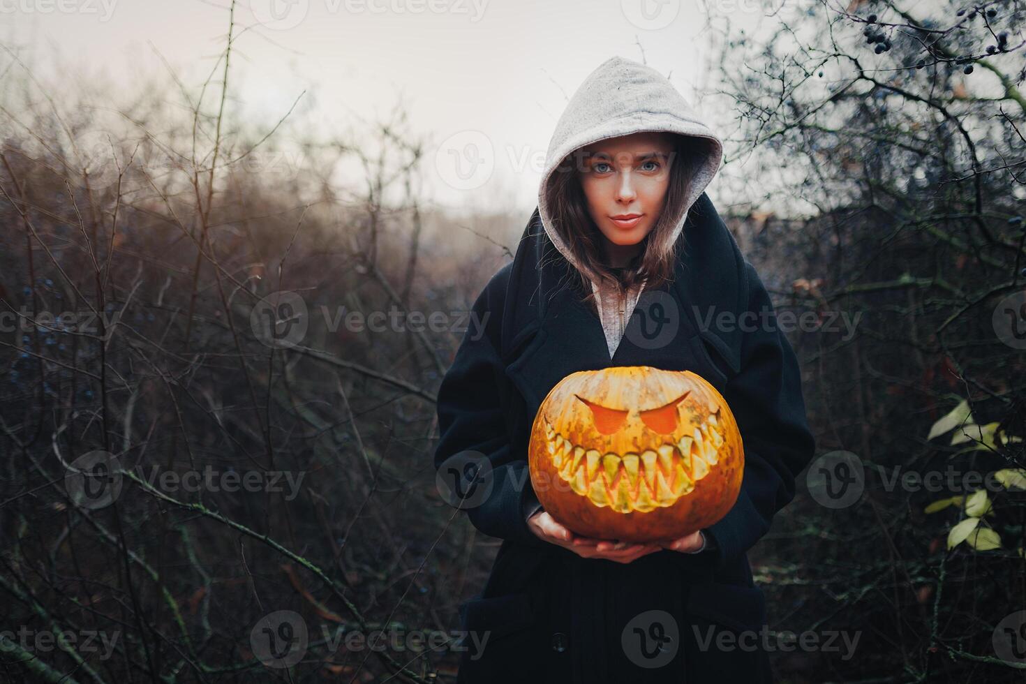 Happy Halloween Portrait of black modern lifestyle witch in hood and black coat mantle on wooden bush forest background. Holding pumpkin in hands photo