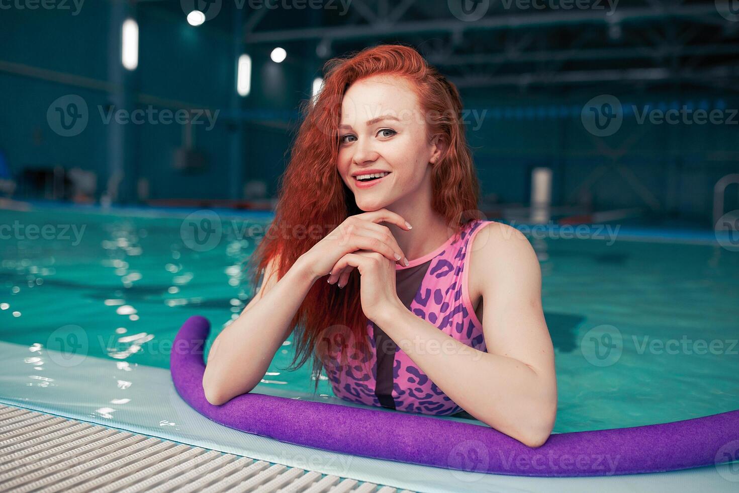 hermosa joven Pelirrojo niña en un de moda traje de baño en el estilo de el Años 80 soportes con el tallarines en su manos en el interior nadando piscina foto