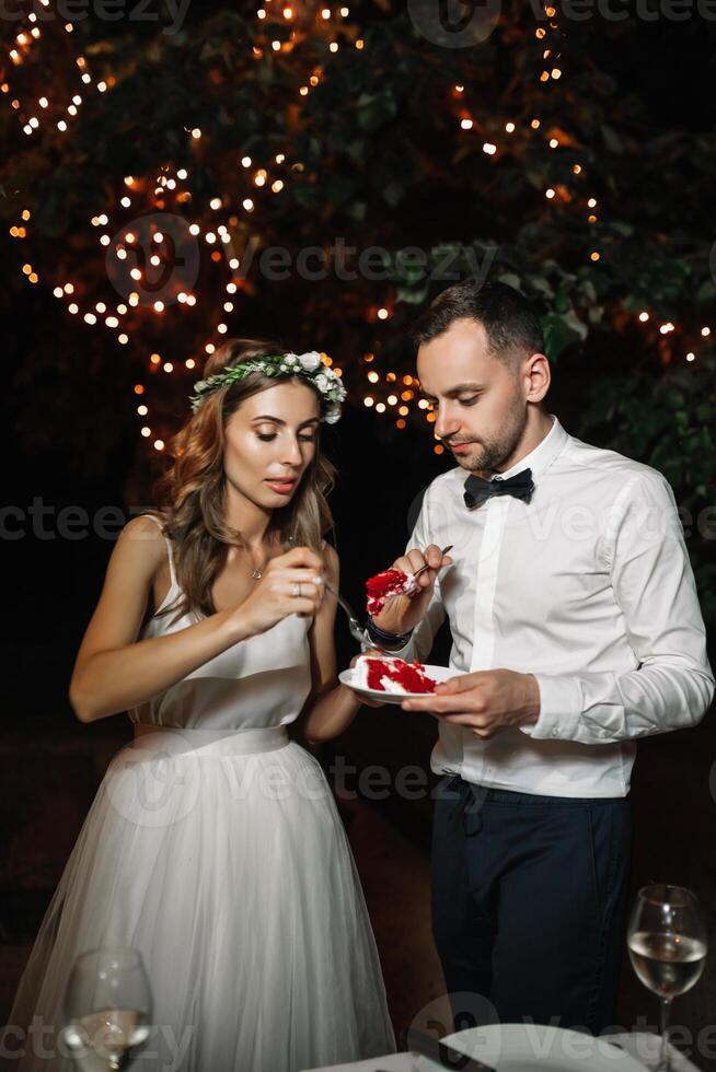 Happy bride and groom cut the wedding cake in the front garland light decoration. Wedding evening outdoor. Sweet happy life concept photo