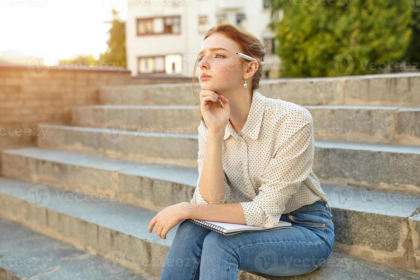 young beautiful student writes an essay in her notebook sitting on the steps stairs outdoor Red-haired girl with freckles photo