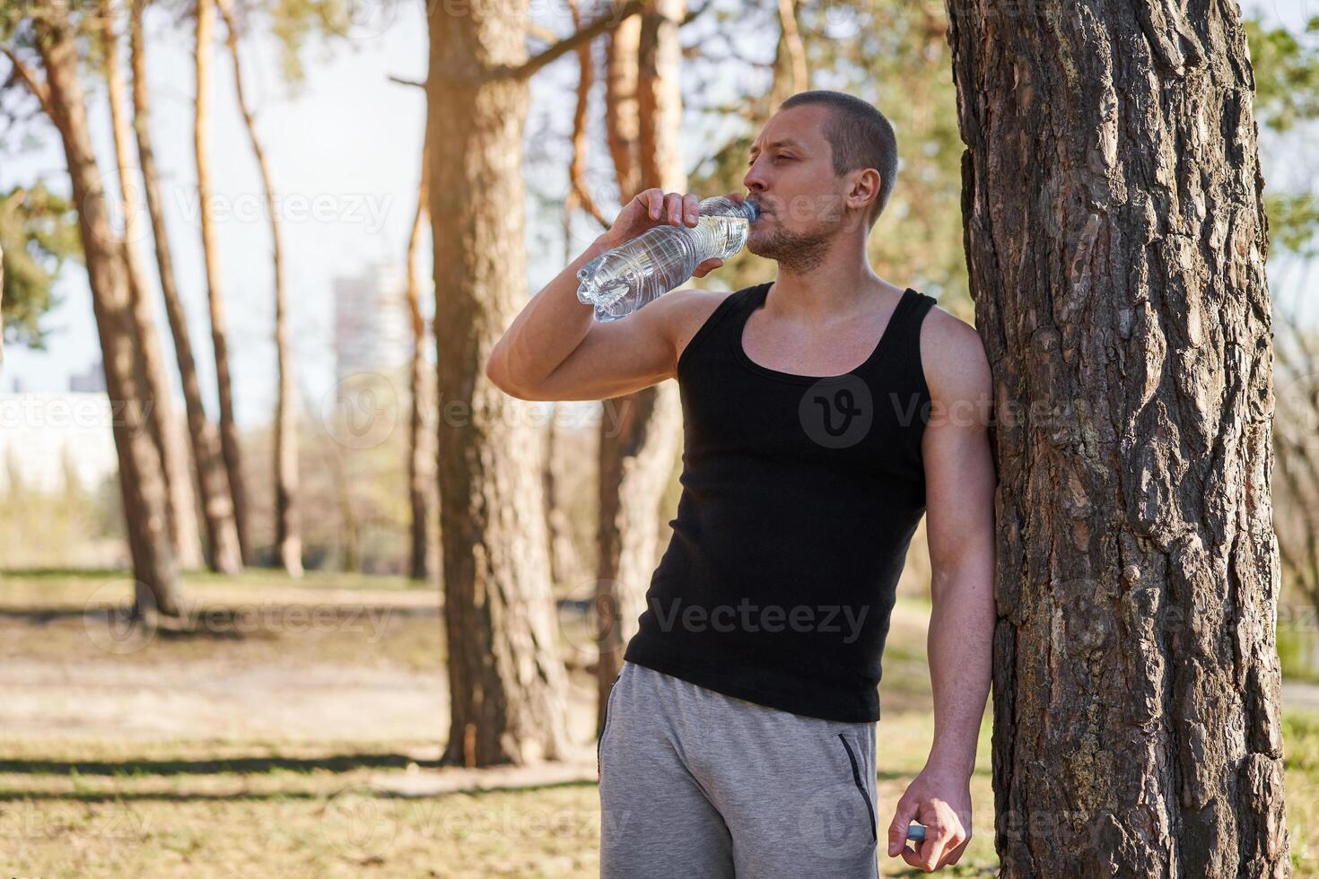 Athlete man drink water plastic bottle workout leaned on tree forest photo