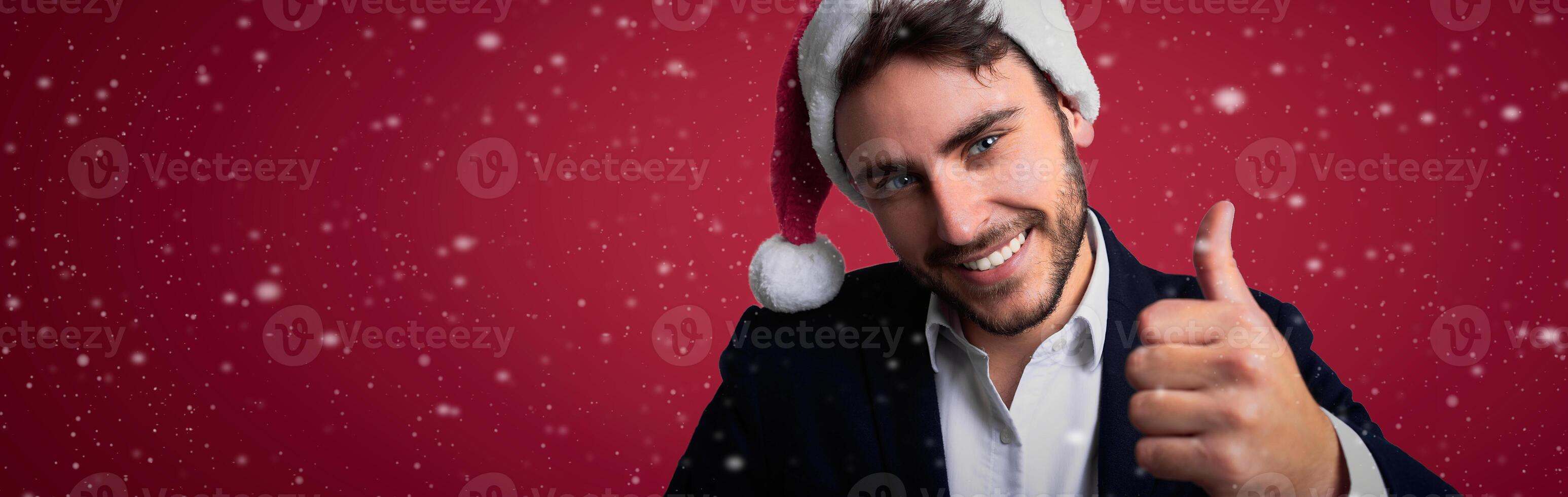 Young handsome caucasian guy in business suit and Santa hats stands on red background in studio smilie and showing thumbs up. photo