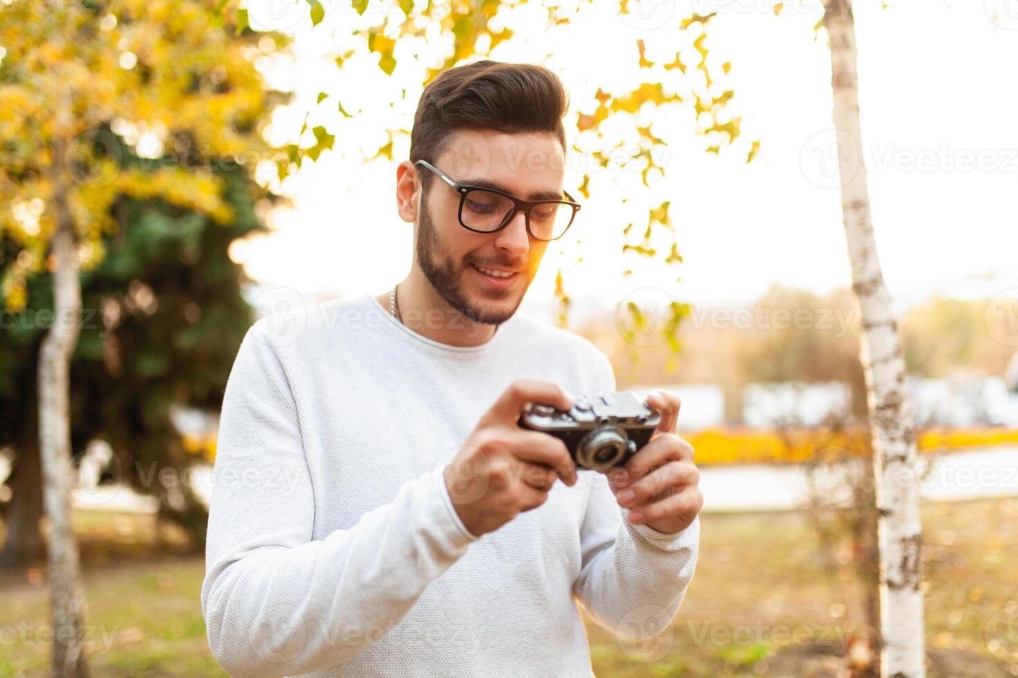 Young handsome hipster guy walks in a beautiful autumn park on the background of yellow leaves in warm sunny weather and takes pictures on a pig film camera. Autumn leisure time. Creative youth photo