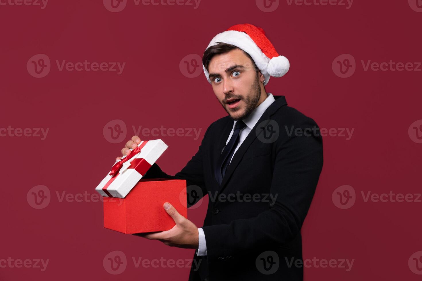 Young handsome caucasian guy in business suit and Santa hats stands on red background in studio and teeth smilie Holding red gift box in hand. photo