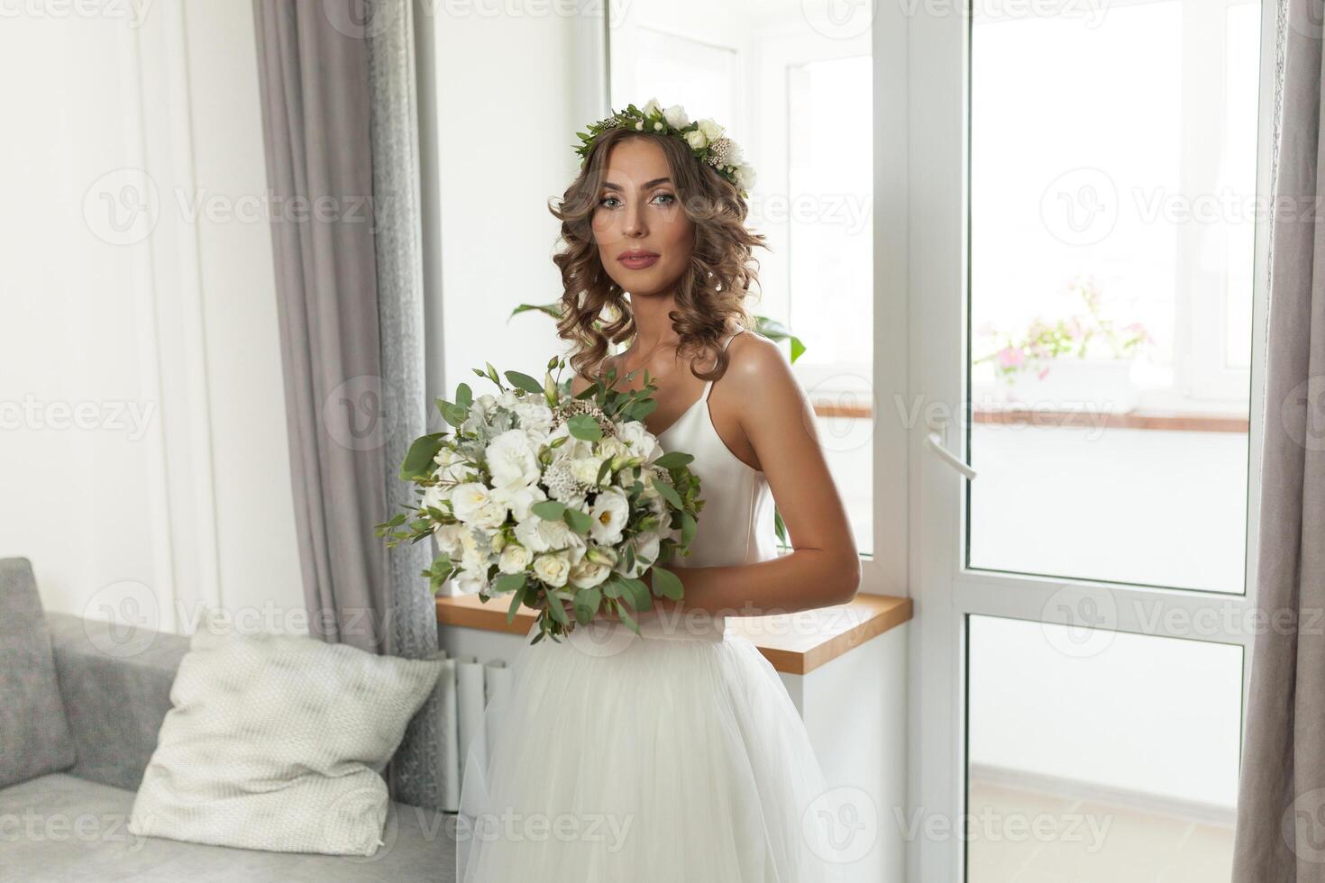 The bride in a beautiful wedding dress with a long curly hair stands in the bedroom and looks at the camera. Bride indoors with white flowers bouquet and tender wreath hairstyle photo
