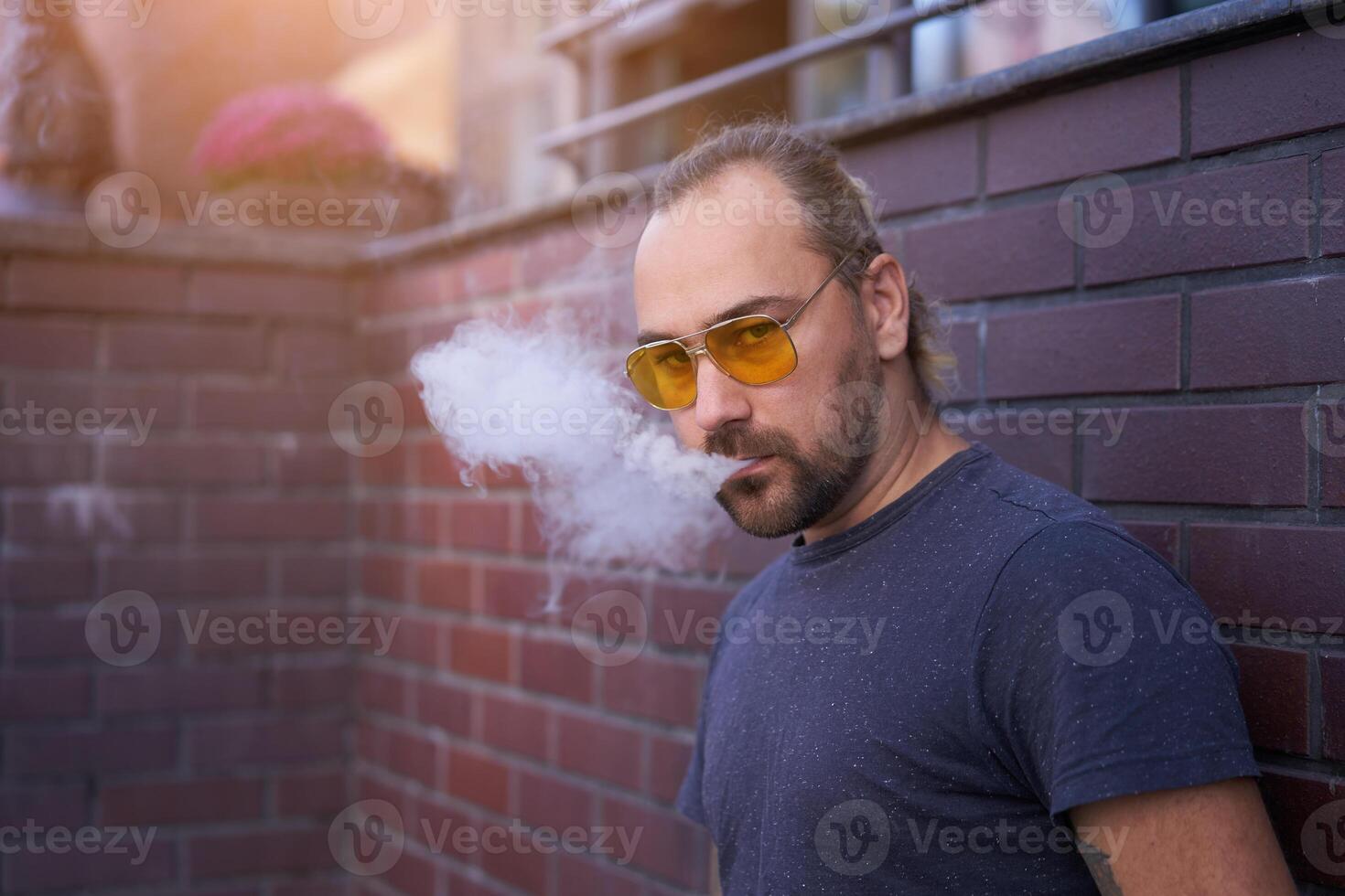 Handsome young unshaven man blue shirt yellow sunglasses stands backyard near brick walls summer day. photo