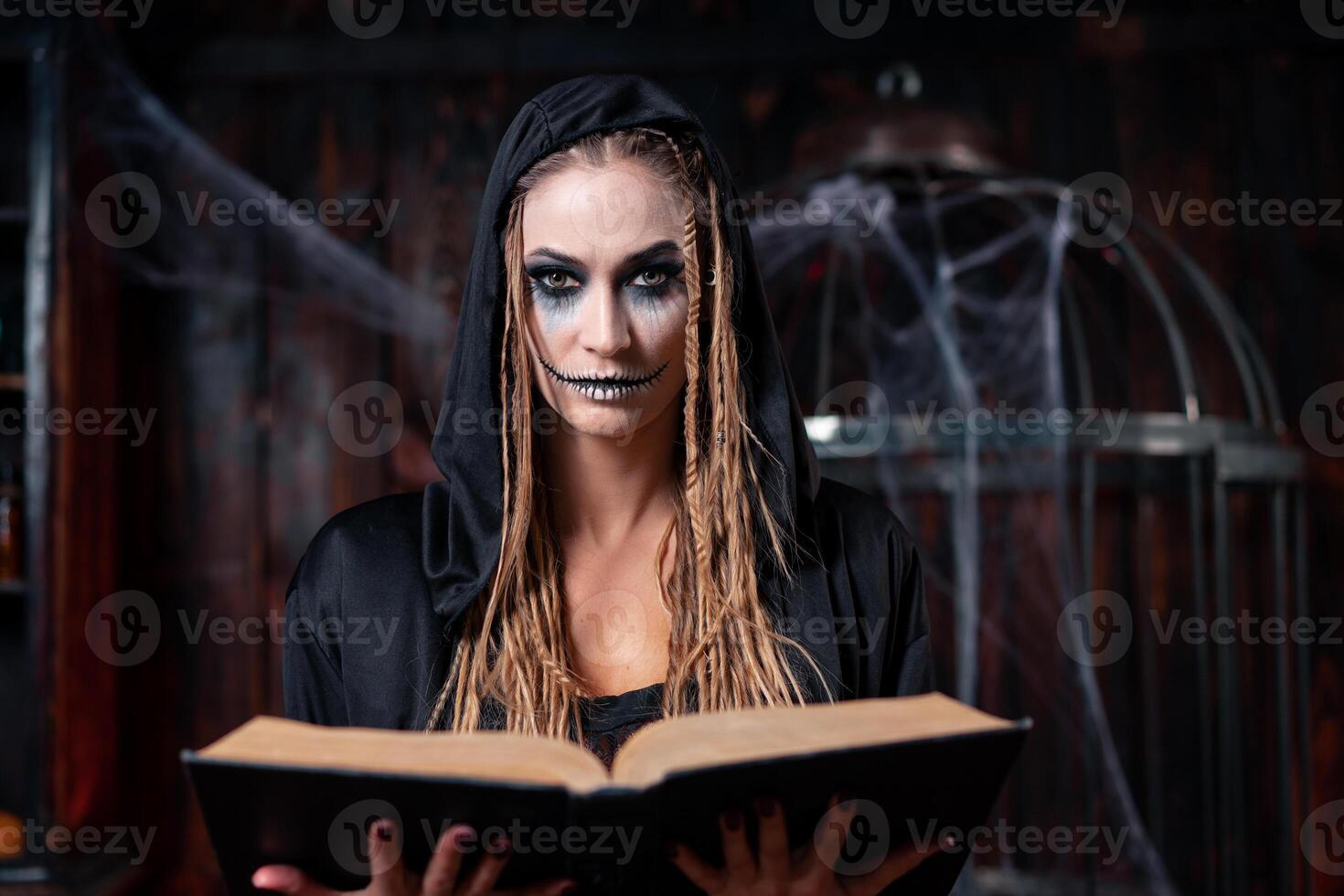 Halloween concept. Witch dressed black hood with dreadlocks standing dark dungeon room use magic book for conjuring magic spell photo