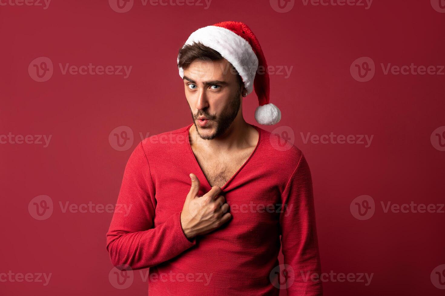 Young handsome caucasian guy in red sweater and Santa hats stands on red background in studio  Shows how hot he is photo