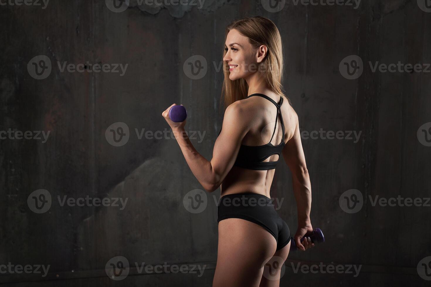 Image of muscular young female athlete wearing black sport wear stands with his back to the camera on dark background. photo