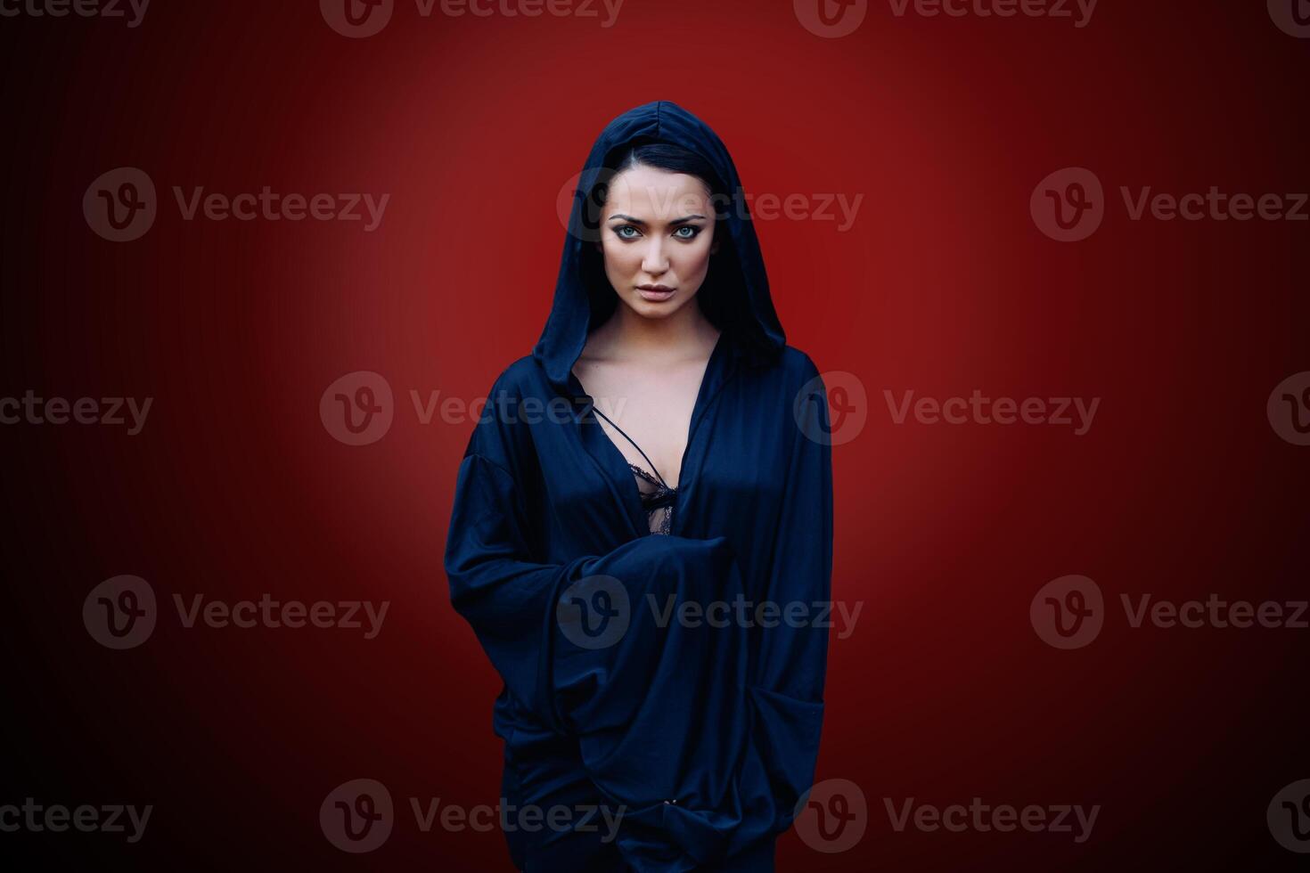 Young beautiful woman with a black hair and in the dark blue cloak with hood at the red background photo