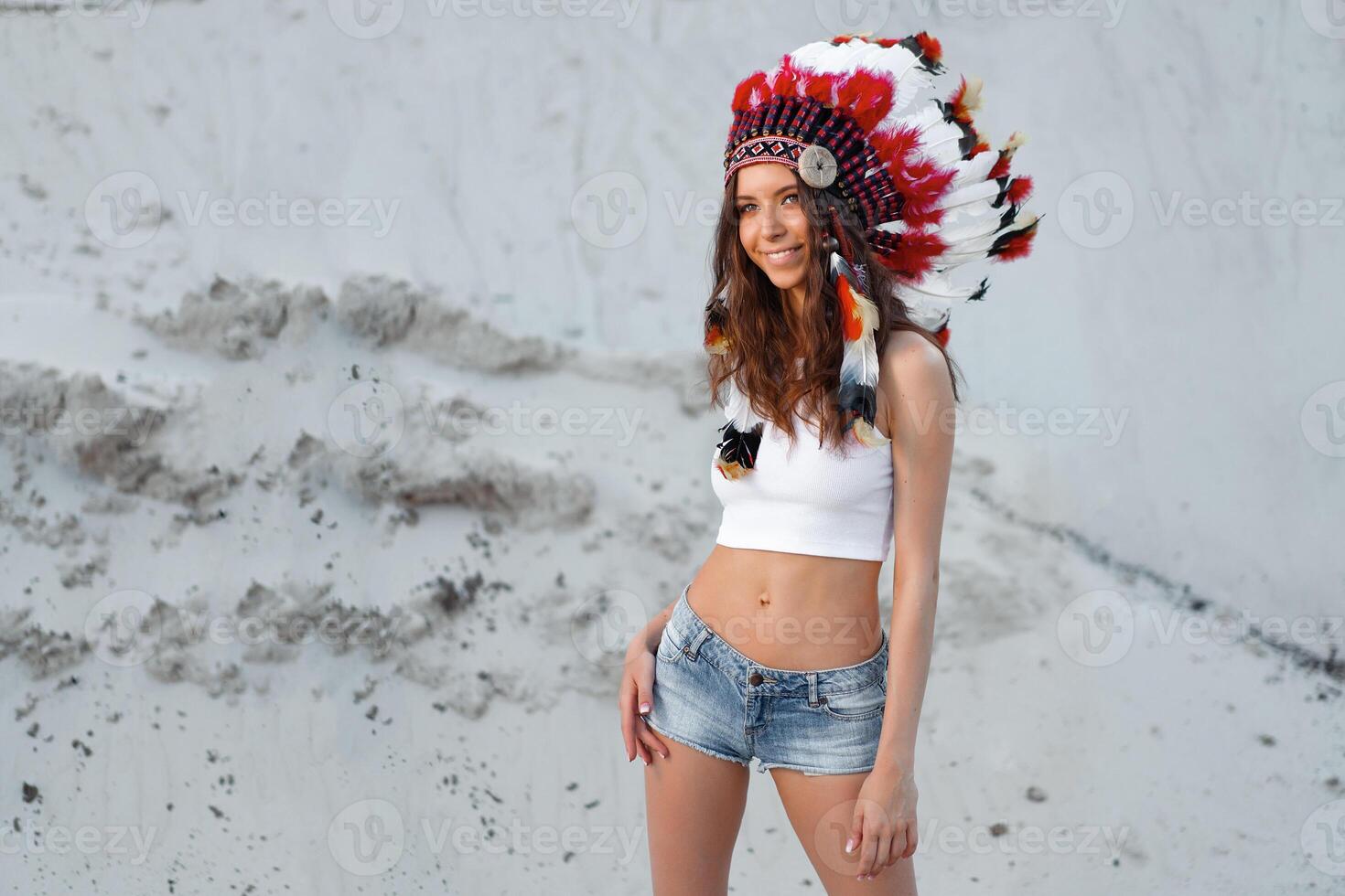 A beautiful young Caucasian girl in a white top and denim shorts on her head is wearing an Indian hat. Roach is in the desert. Happy holiday mood. photo