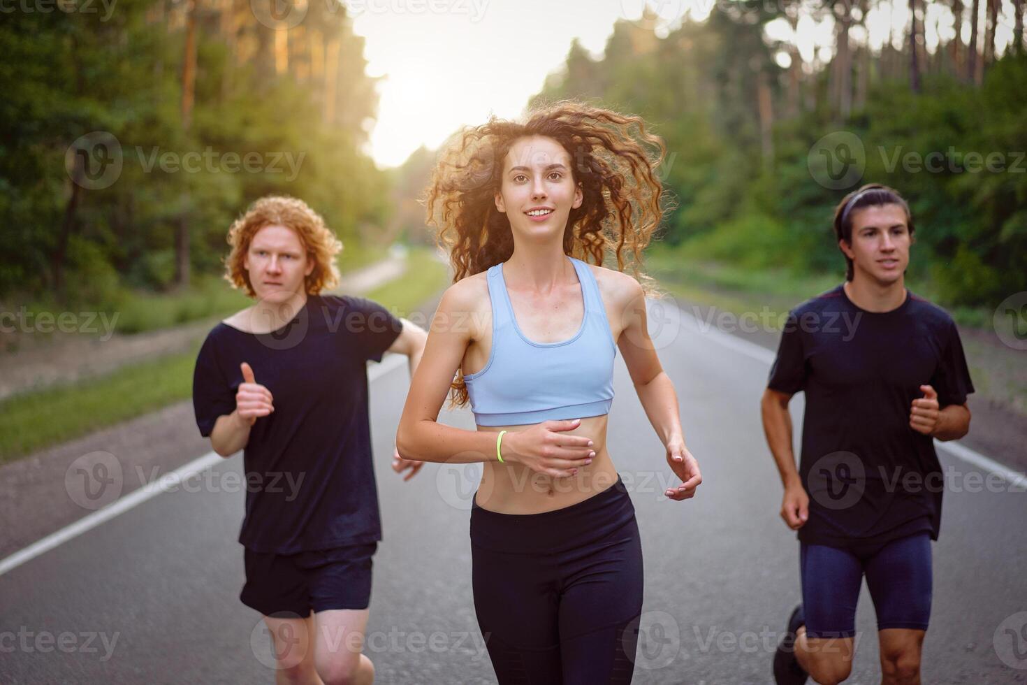 A group of three people athletes one girl and two men run on an asphalt road in a pine forest. photo