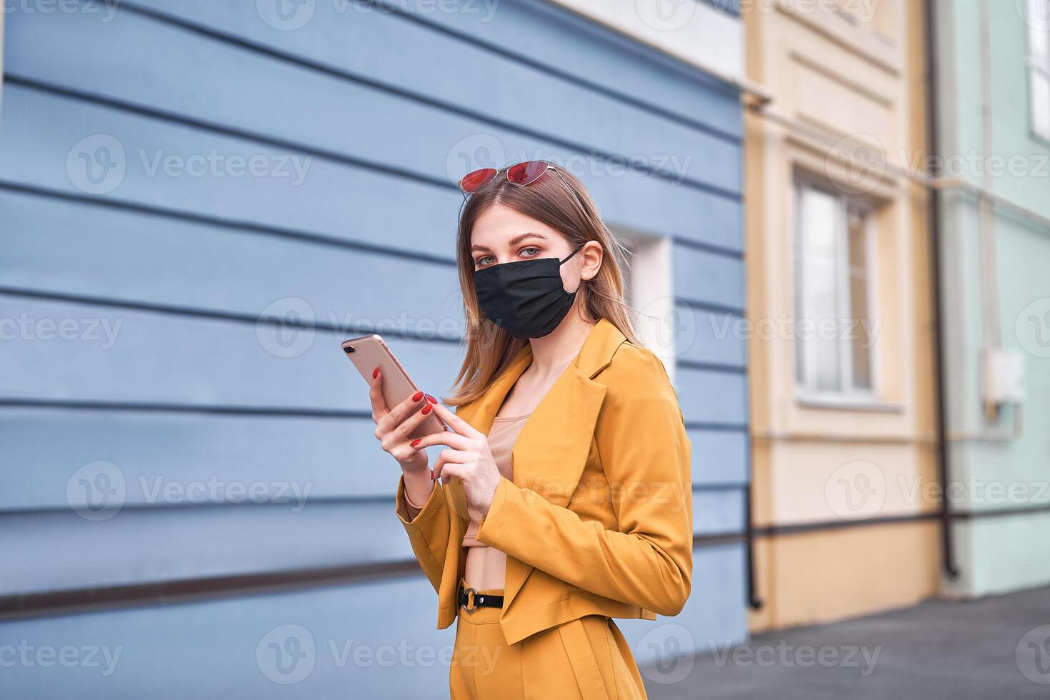 joven hermosa caucásico niña 20 años vestir negro cara máscara proteccion en contra epidemia coronavirus covid-19 camina abajo el calle mira a el teléfono pantalla. foto