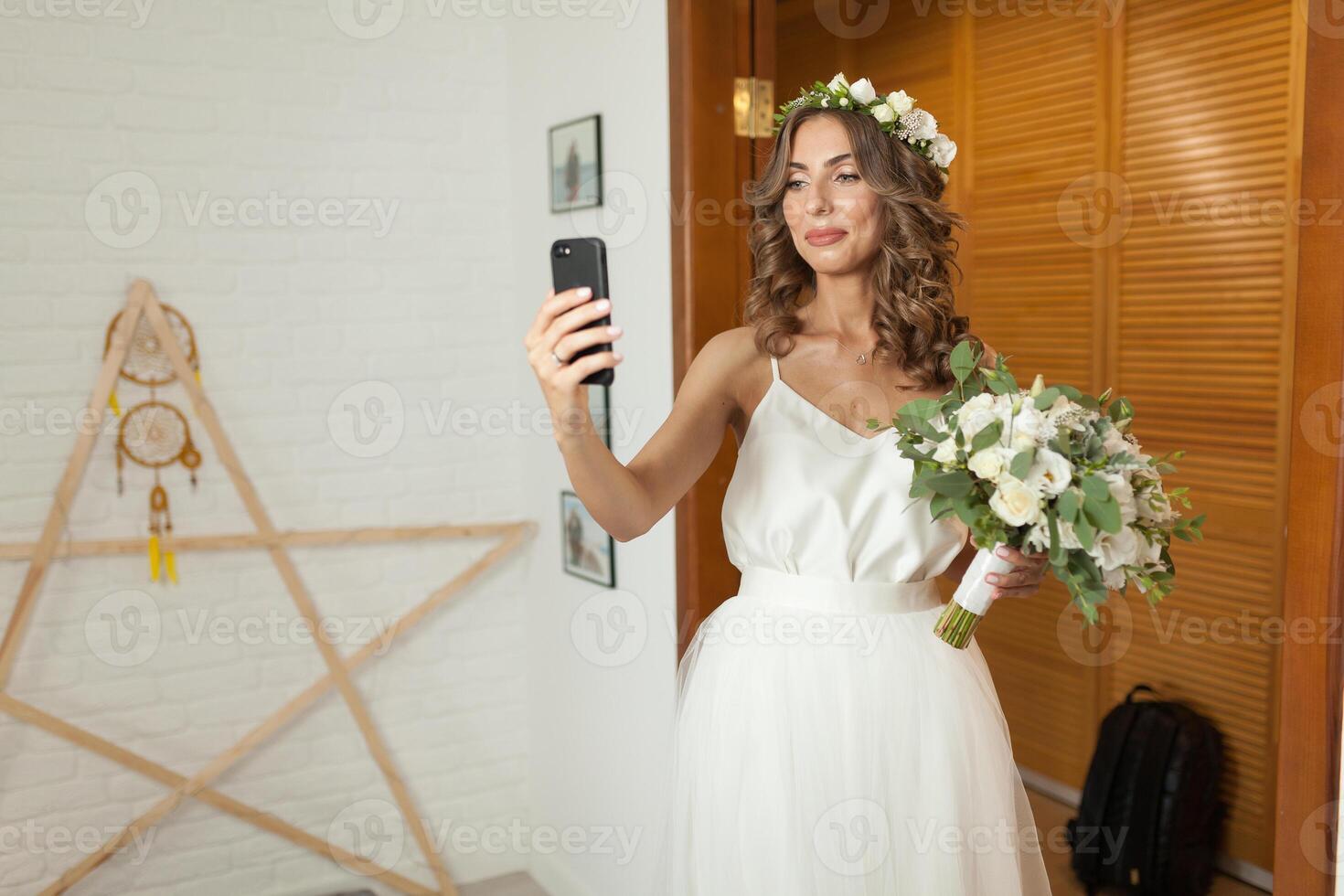 Romantic and happy caucasian bride in stylish wedding dress taking selfie on the background of beautiful room. Taking the bouquet in hand photo