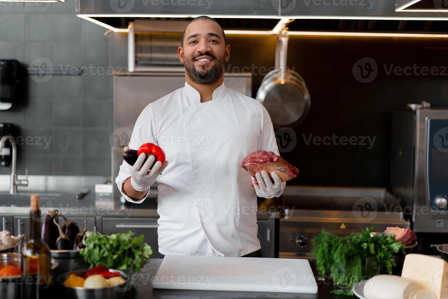 Happy smiling chef prepares meat dish with various vegetables in the kitchen. In one hand the man holds vegetables, in the other a piece of fresh meat photo