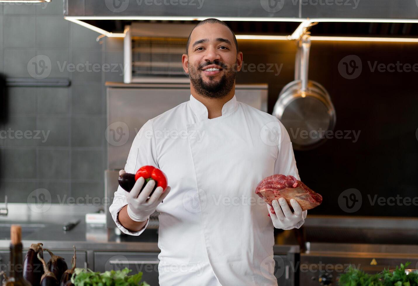 Happy smiling chef prepares meat dish with various vegetables in the kitchen. In one hand the man holds vegetables, in the other a piece of fresh meat photo