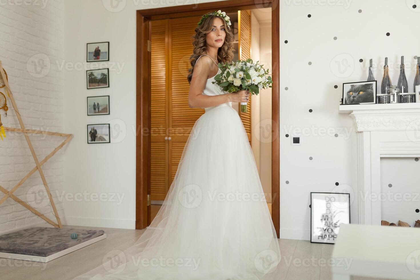 The bride in a beautiful wedding dress with a long curly hair stands in the bedroom and looks at the camera. Bride indoors with white flowers bouquet and tender wreath hairstyle photo