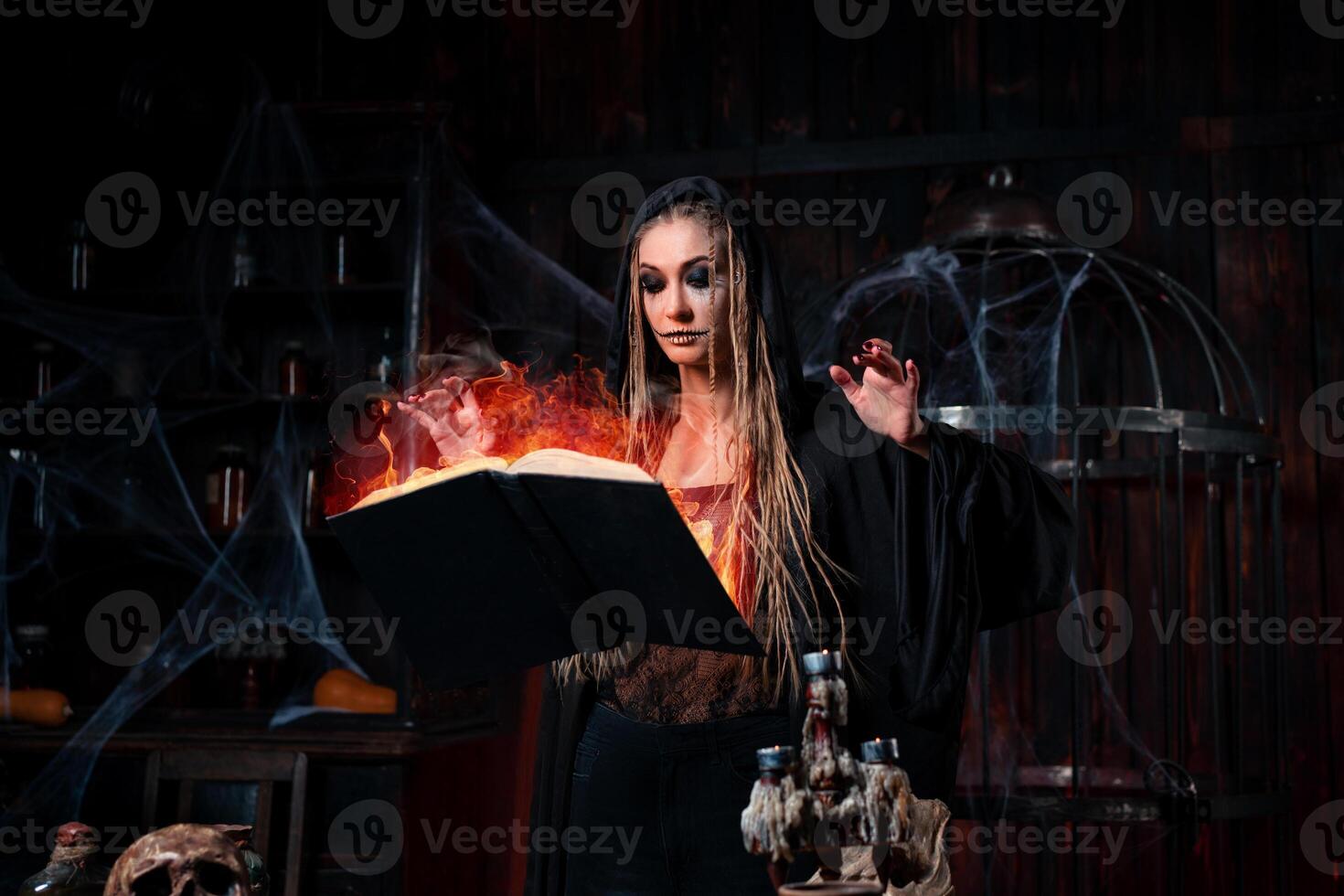 Halloween concept. Witch dressed black hood with dreadlocks standing dark dungeon room use magic book for conjuring magic spell photo