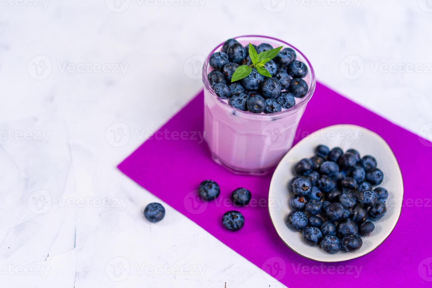 Tasty fresh blueberry yoghurt shake dessert in glass standing on white table purple napkin background. photo