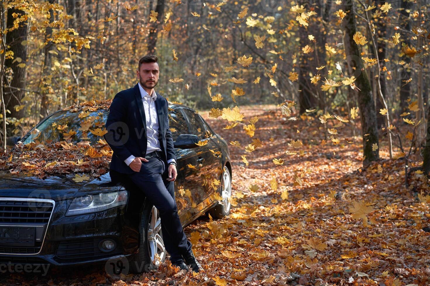 A handsome young Caucasian man wearing business suit standing near black car on nature. photo