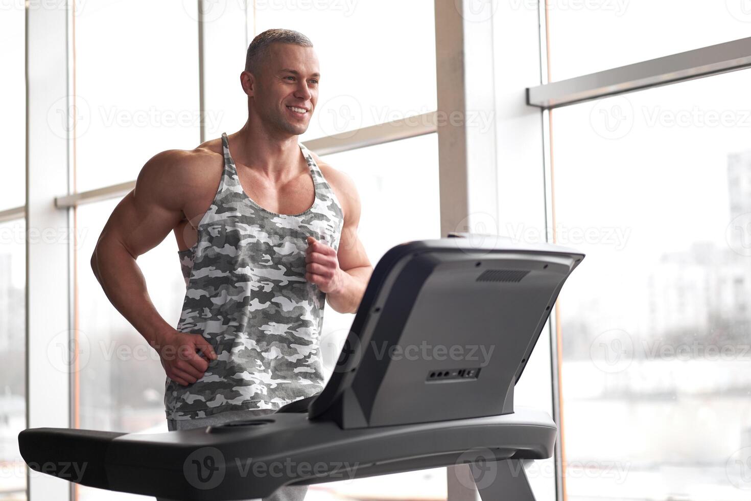 Muscular athletic bodybuilder fitness model running treadmill gym near big window photo