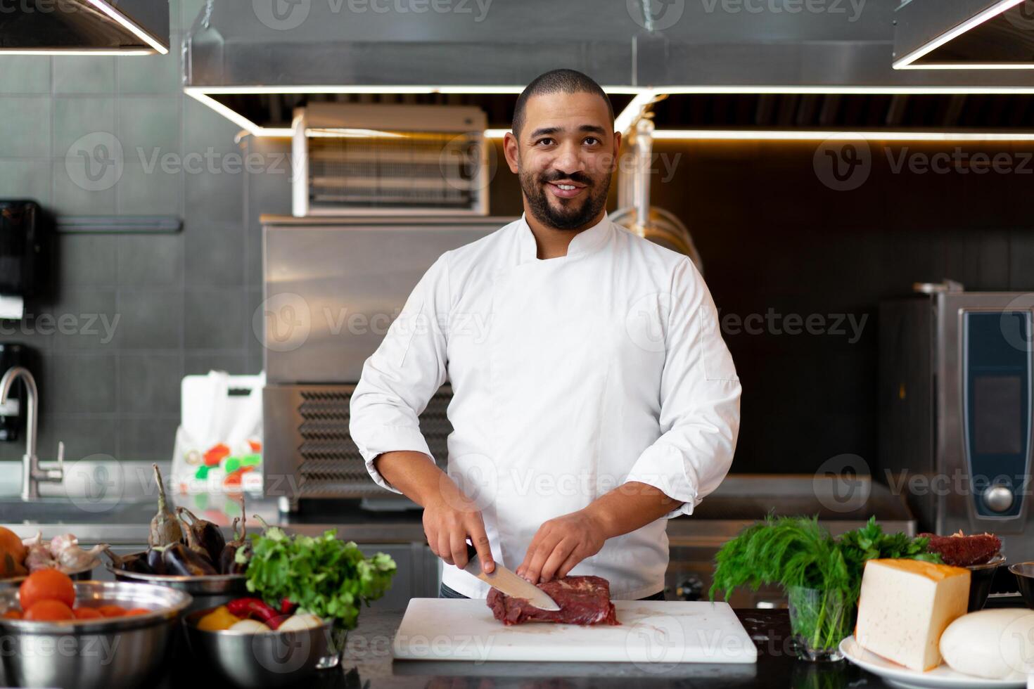 Handsome young African chef standing in professional kitchen in restaurant preparing a meal of meat and cheese vegetables. photo