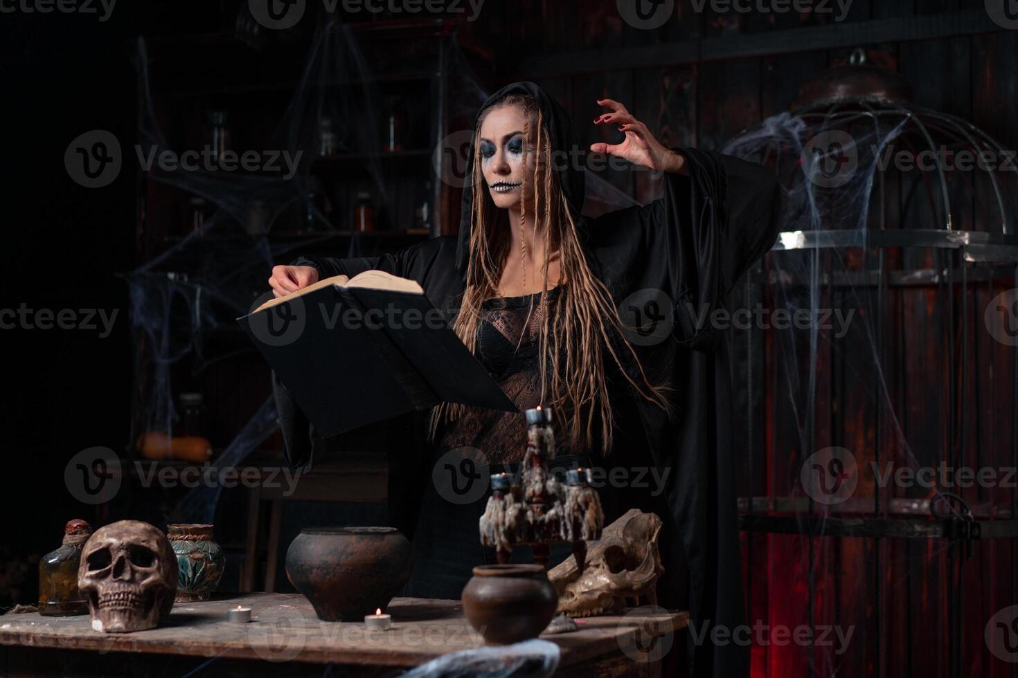 Halloween concept. Witch dressed black hood with dreadlocks standing dark dungeon room use magic book for conjuring magic spell photo