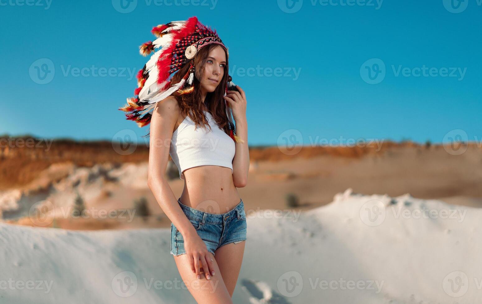 A beautiful young Caucasian girl in a white top and denim shorts on her head is wearing an Indian hat. Roach is in the desert. Happy holiday mood. photo