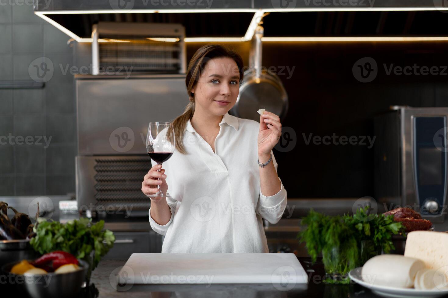 Beautiful young caucasian girl standing in kitchen in a white uniform smiling and tasting red wine photo