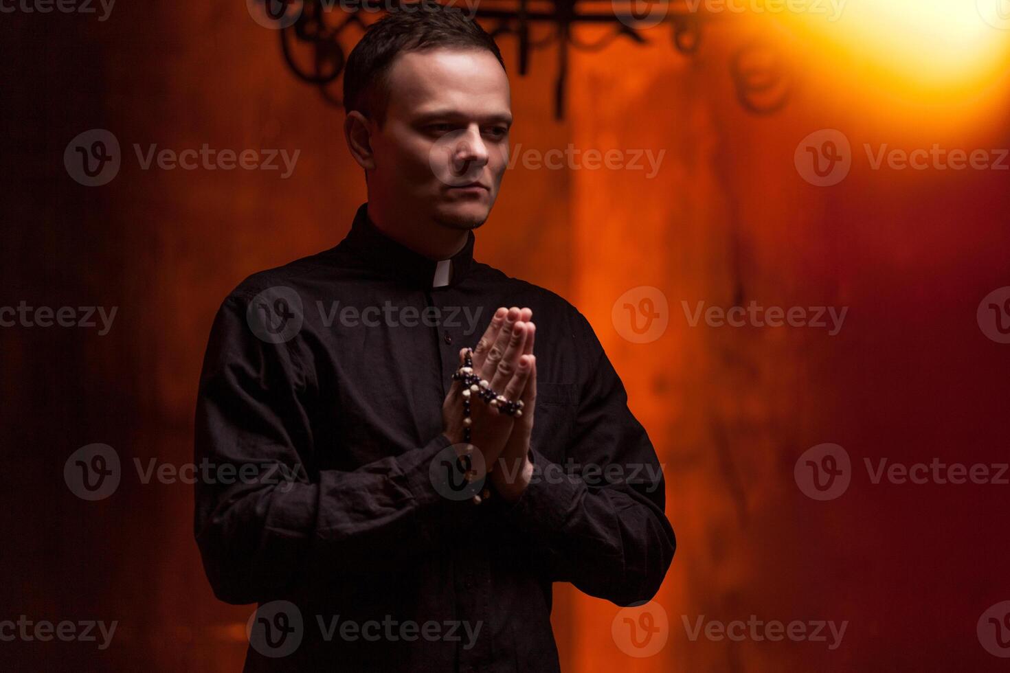 Young Catholic Praying priest. Portrait of priest Next to the candles prays with his hands folded near the face. Red wall on the background. Indoor handsome portrait of religion man photo