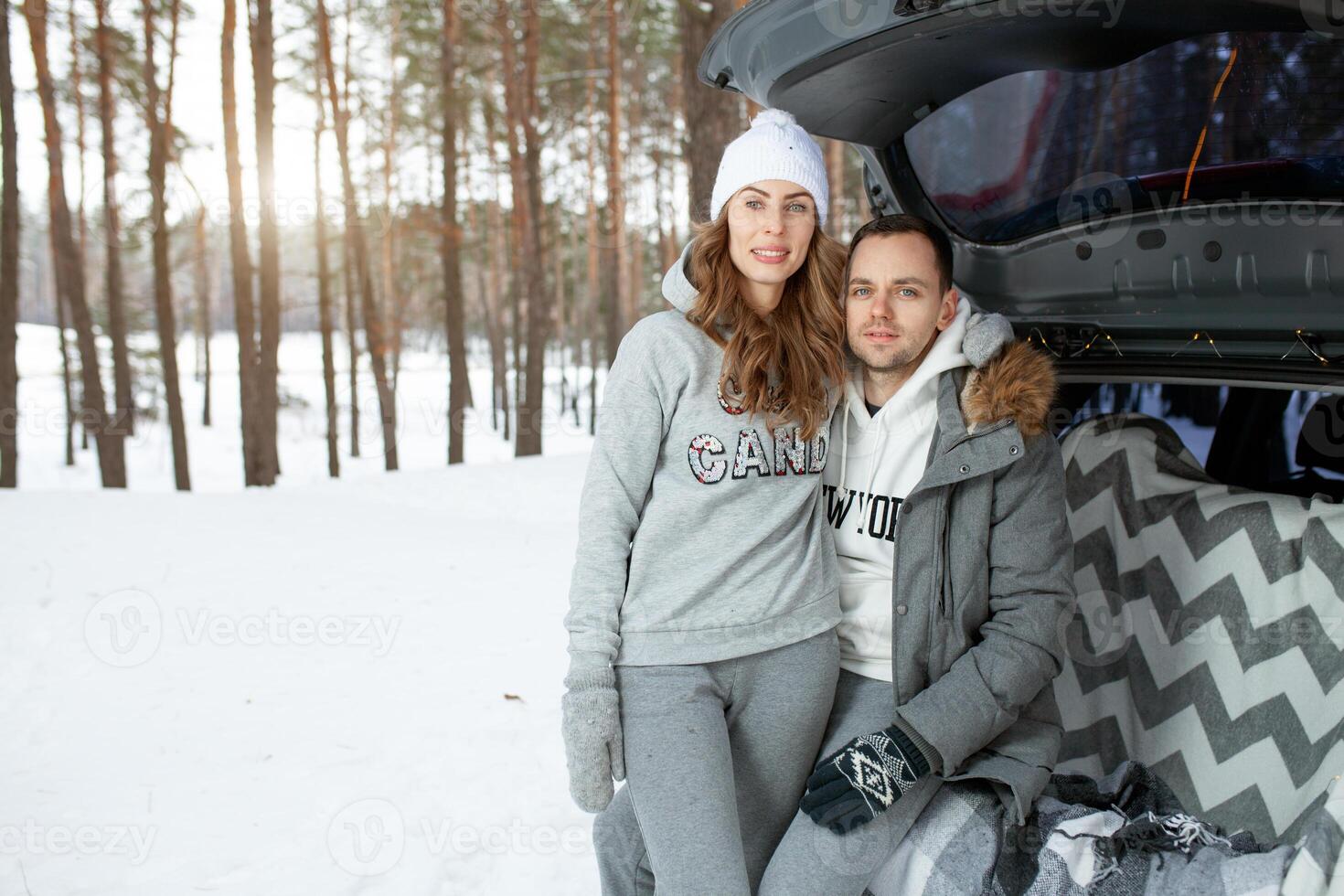 A young family of travelers guy and girl are sitting in the trunk of his car in an embrace. A walk of a young couple in the winter pine forest. Love photo