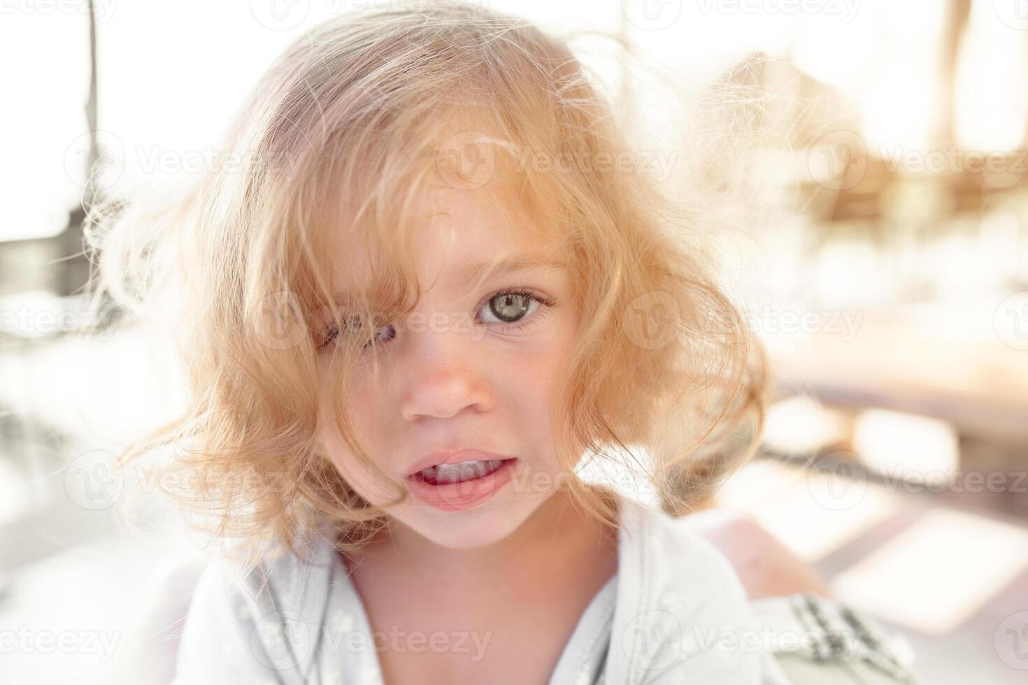 Close-up portrait of pretty young little blond pale unhappy moody friendless child girl looking sadly in camera on blurred sunny outdoors background. Children problems concept. photo
