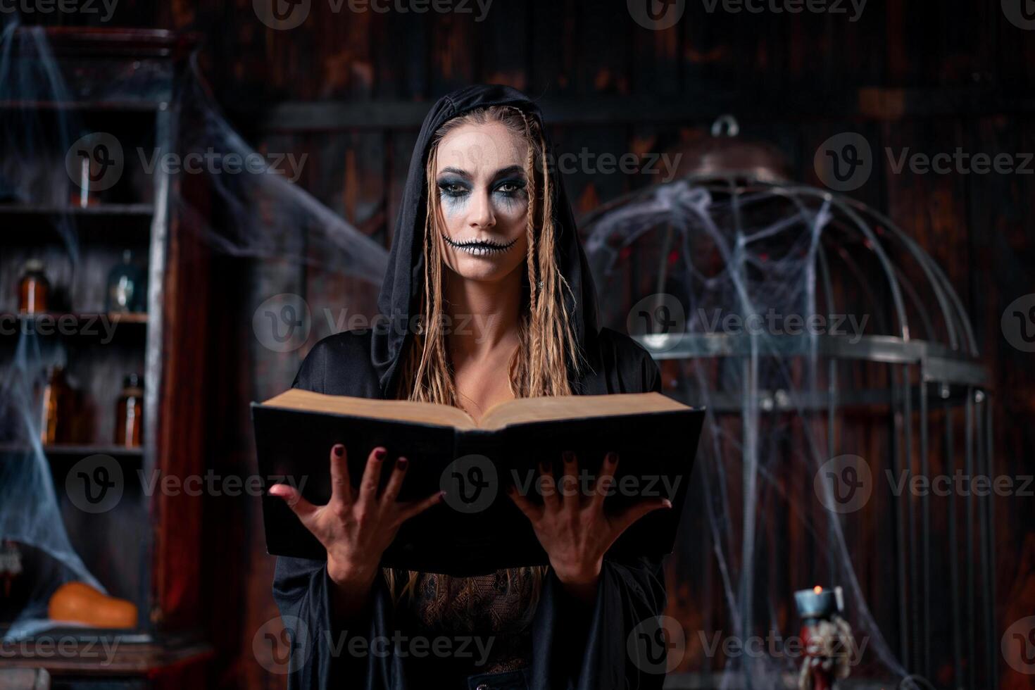 Halloween concept. Witch dressed black hood with dreadlocks standing dark dungeon room use magic book for conjuring magic spell photo