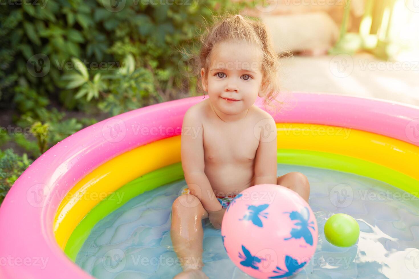 Children playing in inflatable baby pool. Kids swim and splash in colorful garden play center. Happy little girl playing with water toys on hot summer day. Family having fun outdoors in the backyard. photo