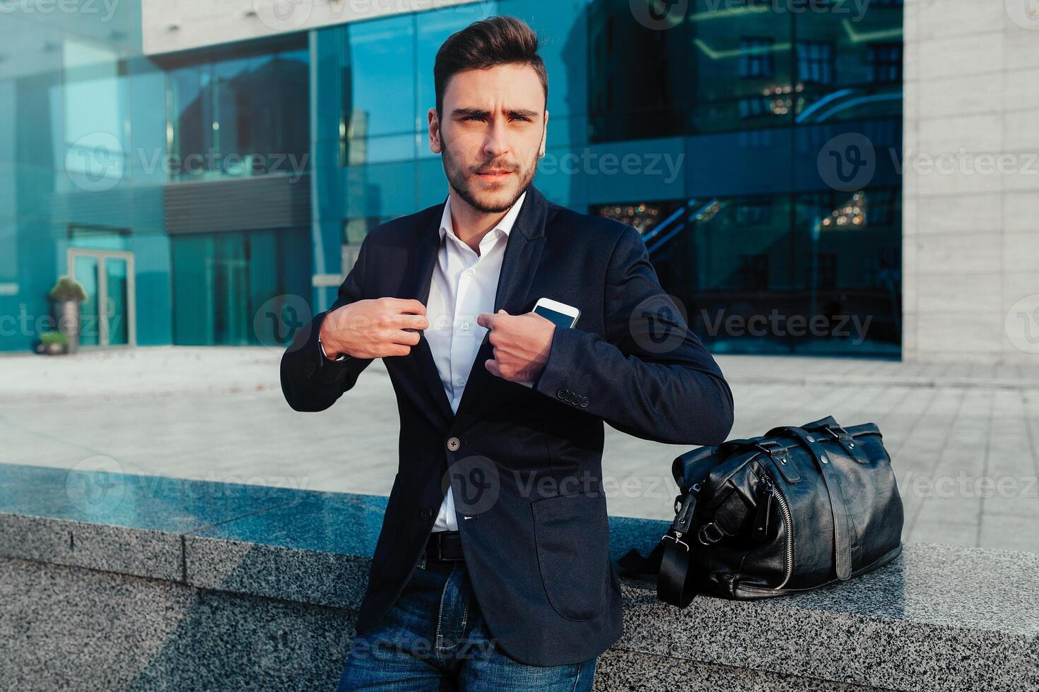 handsome young businessman with a beard and in a business suit standing on the street against the background of the office building next to a comfortable stylish leather bag. photo