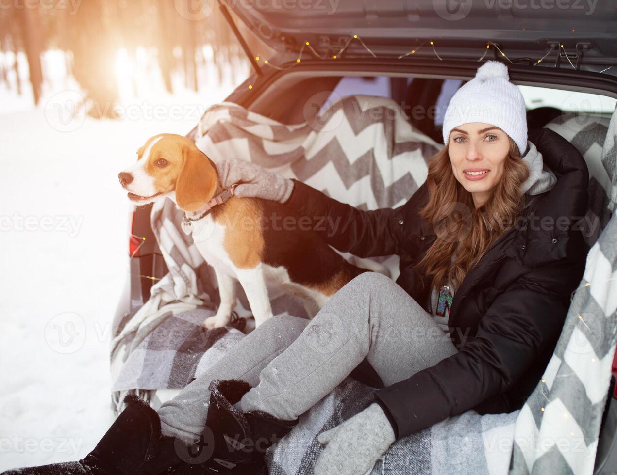 A young family of travelers woman are sitting in the trunk of their car in an embrace with their friend dog Beagle. Winter forest on the background. Traveling with a dog by car photo