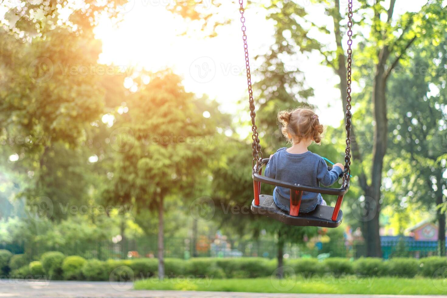 Unrecognizable Little Caucasian girl riding swing at playground sunny summer day rear view photo